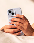 A close-up of a person holding a light blue smartphone case with both hands, resting on soft cream-colored bedding under warm natural sunlight.