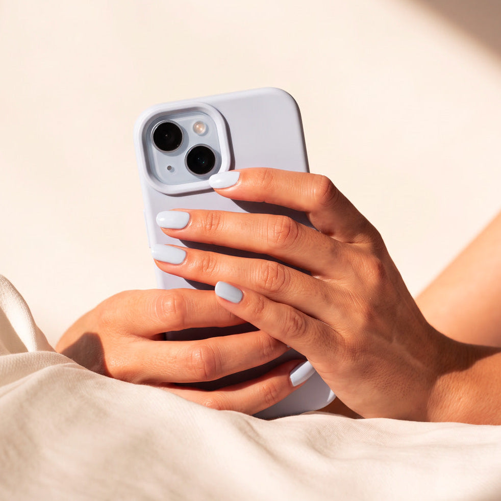A close-up of a person holding a light blue smartphone case with both hands, resting on soft cream-colored bedding under warm natural sunlight.